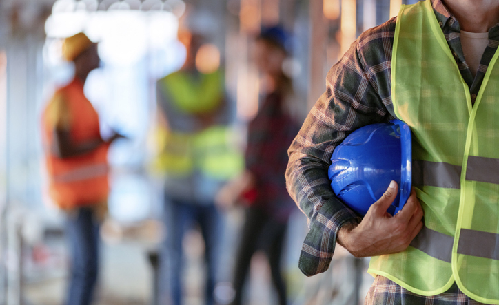 Man holding yellow helmet close up. Construction man worker with office and people in background. Close up of a construction worker's hand holding working helmet.