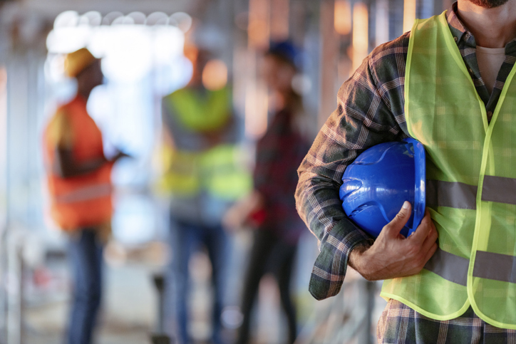 Man holding yellow helmet close up. Construction man worker with office and people in background. Close up of a construction worker's hand holding working helmet.