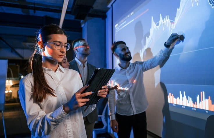 Woman is standing with tablet. Male leader talking to employees, showing the plan on the projector in office.