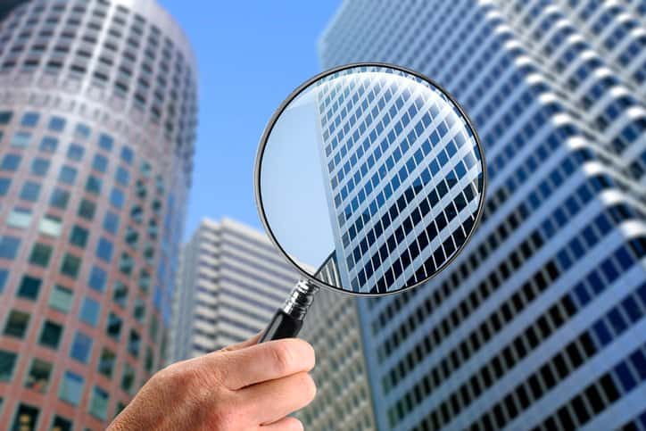 A man holds a magnifying glass in front of a group of highrise office buildings.
